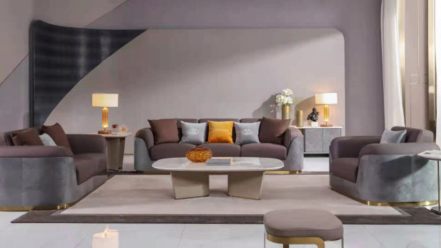 This picture shows a beautiful, modern living room design. The room has a large white marble floor, with a light gray area rug in front of the sofa. The walls and ceiling are a light gray color. There is a large modern gray sectional sofa in the room, along with a contemporary glass top coffee table and two white armchairs. On the wall behind the sofa is an abstract art piece, and the room is accented with decorative pillows, throw blankets, and a few small side tables.