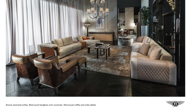 This picture depicts a luxurious and modern living room with an open floorplan. The main focal point of the room is the grey velvet L-shaped couch, which is nestled in a corner and positioned in front of a large flat-screen television mounted to the wall. There are also two gold-accented armchairs nearby. The walls and floors of the room have been painted a neutral gray, with a white ceiling, providing a blank canvas for various artworks, like the metal wall sculptures on either side of the TV. The space is illuminated by a large, metal chandel