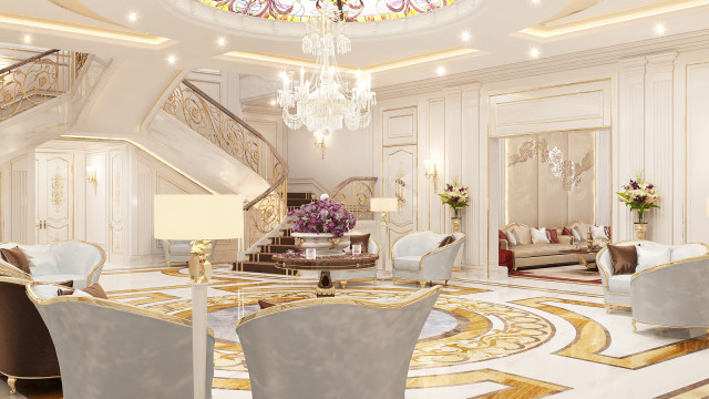 This picture shows a luxurious home interior. The room has an elegant marble floor, with a gold and white patterned rug in the center. There is also a large sofa with plush cushions in a beige color, and a decorative fireplace with intricate detailing. The walls are painted in a light gray shade, and there are several pieces of artwork hung up on them. There is also a grand staircase in the center of the room, with a chandelier above it.