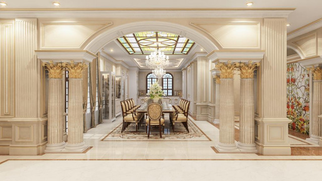 This image is of a luxurious living room designed by Antonovich Design. It features a custom arch ceiling with a patterned design, ornately carved wooden furniture accented with gold leaf details, white marble flooring, and a floor to ceiling glass window with a forest landscape outside. The room is illuminated by grand crystal chandeliers and other light fixtures, while plush rugs and textured fabrics provide an added touch of elegance to the space.