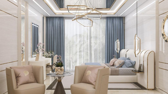 This picture shows a luxurious and modern living room from the Antonovich Design Company. The room has a grey and white color palette with accents of black, gold, and blush pink. A large chandelier hangs from the ceiling and two comfortable couches are arranged around a classic marble coffee table. The area rug in the center of the room adds texture and warmth, while the sleek fireplace and media console create an elegant focal point.