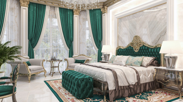 This picture shows a luxurious bedroom decorated with contemporary design elements. The walls are covered in a textured light grey, and the floors are covered in a tailored beige carpet. The bed is upholstered in a white velvet material, and the headboard features intricate diamond stitching. On either side of the bed are matching side tables with gold accents and glass tops. A large velvet armchair is situated in the corner, and several decorative items such as pillows and lamps are scattered around the room.