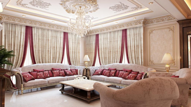 This picture shows a luxury living room set up in a modern style. The room features a large cream-colored sofa with two pillows and an ornate, white armchair with two red cushions and a matching ottoman. To the side of the seating is a large gold-framed portrait and an ornate, faux marble table with a golden lamp and two figurines on top. The walls of the room are painted with a deep aqua hue and feature several intricate sconces with cream-colored shades. A pale brown rug with a geometric pattern stretches across the room