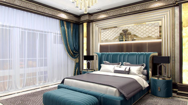 This picture is of a luxurious residential interior design. It features a grand marble staircase, a reception area with white walls, a blue tufted velvet sofa, an ornate chandelier, and an ivory-colored ottoman. The space is complemented by abstract artwork, a classical white clock, and gold-rimmed mirrors. The overall aesthetic is classic and opulent.