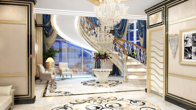 This picture depicts a luxurious living room with plush cream-colored furniture. The walls and ceiling are painted in a deep blue shade, and the floors are tiled in white marble. A large mirror is situated above a sophisticated fireplace, and a crystal chandelier hangs from the ceiling, illuminating the room. There is a grand piano in the corner, and numerous pieces of artwork adorn the walls. A grand entranceway lined with tall columns leads off to another room or area.