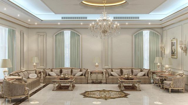 This picture shows a luxurious interior design with an ornate balcony overlooking a large room. The room features a grand chandelier in the center with cream-colored walls and classic furniture pieces. A long cream-colored sofa stretches along the length of the room, and two white armchairs sit on either side of a warmly-lit fireplace. On the far wall, an elegant, white lace drapery frames an ornate mirror.