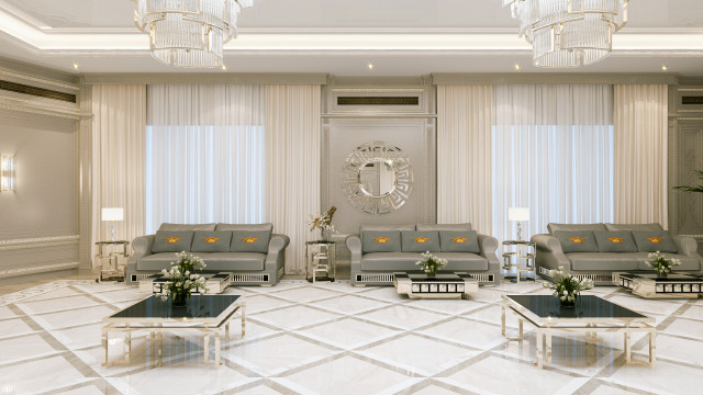 This picture shows an elegant living room in a modern style. The room features sleek light grey walls, white trim and furniture, and a large crystal chandelier for a touch of glamour. The floor is covered in a plush grey rug, and the seating includes a cream velvet sofa and two beige armchairs with an accent table between them. There is also an elegant white marble fireplace with a black mirror hanging above it.