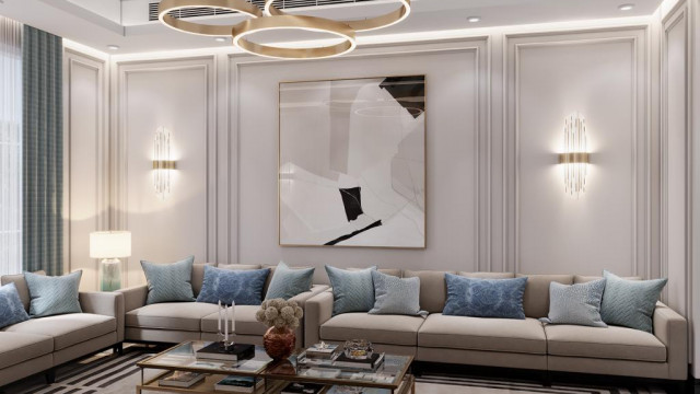 The picture shows a modern, luxurious living room designed by Antonovich Design. The room is decorated in tones of beige and cream, with a plush rug spanning the length of the room. The walls are lined with recessed alcoves which display art pieces. The seating area consists of two off-white couches and two armchairs upholstered in beige velvet. The space is framed by two large windows with panels of sheer fabric hung at either side.