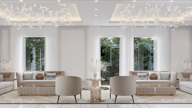 This picture shows an interior design of a modern living room. The room has a large sectional sofa and two armchairs upholstered in beige fabric. There is a white coffee table in the center, and a white and grey rug covers the floor. Large windows line one wall, letting in natural light and offering views onto the garden outside. The walls are painted in a light grey shade and feature artwork and shelves.