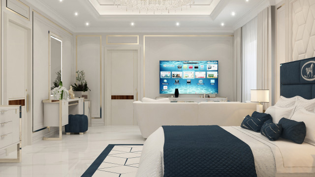 This picture shows a luxurious living room with a curved white sofa, an ornate chandelier, and a large glass window. The walls are decorated with intricate gold detailing and there is a rug with a floral pattern on the floor. A white table sits in the corner with a vase of flowers, adding a touch of elegance to the room. The overall design is both modern and classy, creating a perfect atmosphere for relaxing and entertaining.