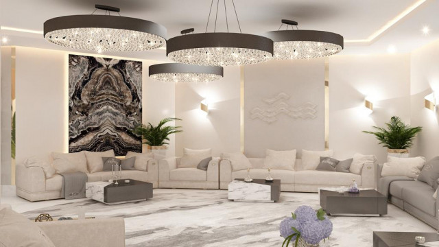 This picture shows a luxurious living room in an opulent modern style with a light grey and white color palette. The room is decorated with a stylish sectional sofa, an ornate decorative rug, and a glass top coffee table. The walls are adorned with a large abstract art piece and accent lighting fixtures, while the dark hardwood floors add a touch of warmth to the space.