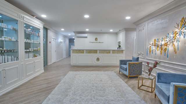 This picture is of an elegant entryway with a sweeping marble staircase. The walls are decorated in a classic style with framed art and ornamental moldings. At the top of the stairs are two large chandeliers, and on the landing below is a polished wood table with an intricate floral centerpiece. There is also a set of doors leading into a grand room beyond.