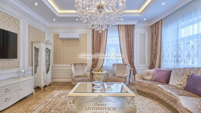 In this picture, we can see a modern and luxurious interior design by Antonovich Design. It includes a sophisticated and stylish living room with white walls and golden details. In addition, the furniture is upholstered in lavish fabrics such as velvet, leather, and fur. There is a plush white sofa with two accent armchairs, and a round marble coffee table. The walls feature abstract art pieces as well as framed photos and mirrors. This beautiful living space also has a round chandelier that adds a stunning and glamorous finish to the design.