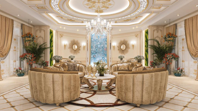 This picture shows a luxurious master bedroom designed by Antonovich Design. The room is decorated with elaborate white and gold accents, giving it a regal and opulent feel. There is a large, tufted headboard on the bed and plush throw pillows in various colors and fabrics. The walls are a light brown and beige color, while the floor is covered in a textured, gold area rug. A crystal chandelier hangs from the ceiling and a tray of champagne glasses sits atop an intricate nightstand.