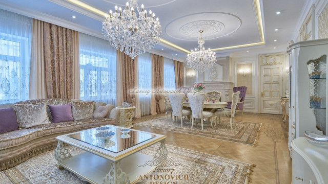 This picture shows a luxurious living room with an abundance of rich details. The walls are covered in a creamy beige and champagne wallpaper, while the ceiling is painted white and adorned with intricate stucco details. There is a large sofa seating area, featuring deep purple velvet sofas and gold pillows, while there is also a warm wooden coffee table with elegant carved legs in the center of the room. On either side of the seating area are two symmetrical consoles with marble tops and ornate mirrors, along with lavish wall sconces for added illumination.