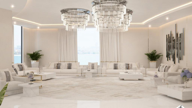 This picture shows a luxurious interior space featuring an ornate chandelier, a grand staircase curved around a marble column, and two crystal sconces on the wall. The walls are painted in a warm peach color, and the floor is detailed with a decorative rug. On either side of the stairs, there are two velvet chairs upholstered in a neutral pattern, providing some cozy seating for the area.