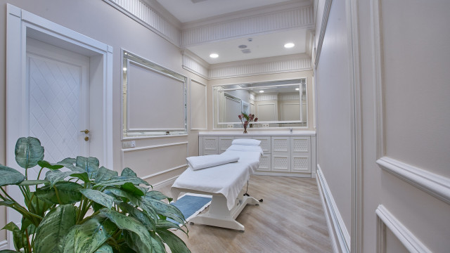 A luxurious bedroom containing a king-sized bed, two nightstands, and a vanity table. The room is decorated with a large crystal chandelier, ornate patterned wallpaper, and classic furniture pieces.