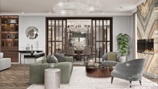 This picture is of a luxurious and elegant dining room interior design. The room contains a large, oval-shaped dining table made of dark wood surrounded by several high-backed chairs upholstered in white fabric. On the wall behind the table is a stunning silver mirror with a decorative frame and two wall sconces framing the top of the mirror. Hanging from the ceiling is a fancy crystal chandelier, adding an extra touch of glamour to the room. The walls are painted a soft grey color with a white chair rail, while a matching rug lies on the floor.