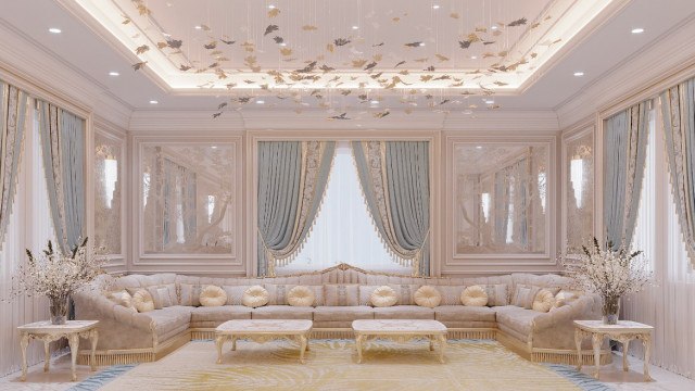 This picture shows a luxurious living room decorated in a classic style. The room has cream colored walls and ceiling, with white crown molding trim. The furniture consists of a beige sofa and chaise lounge, as well as two dark leather chairs and ottomans. The centerpiece of the room is a dramatically large crystal chandelier that hangs above the seating area. There are also decorative pillows and rugs scattered throughout the space that add to the overall classic and elegant feel.