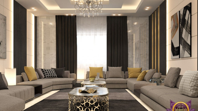 This picture shows a luxurious living room designed by Antonovich Design. The space is decorated with intricate molding and features a grand, grey marble fireplace. The walls are painted in a light cream color and adorned with gold accents, while the furniture is upholstered in plush white fabric and adorned with ornate gold detailing. A large, black chandelier hangs above the space, providing an elegant aesthetic.