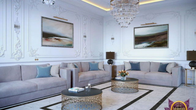 This picture shows a luxury living room designed by Antonovich Design. The room boasts opulent decor and furniture, with a large gold chandelier hanging from the ceiling, cream colored sofas and armchairs, and marble accents throughout. The walls are adorned with intricate patterned wallpaper and there is a plush grey rug covering the floor.