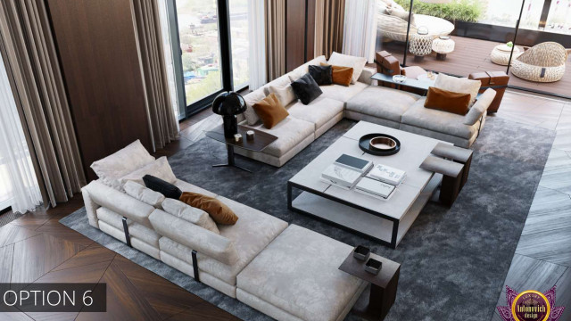 The picture shows a bright and airy living room with white walls and large windows. In the center of the room is a white sofa with a modern, curved design. To the left of the sofa is a large grey armchair, and to the right are two abstract art pieces mounted on the wall. In front of the sofa is a wooden coffee table with a glass top and decorative accessories. Further back in the room is a kitchen area with white cabinetry, a large island, and stainless steel appliances.