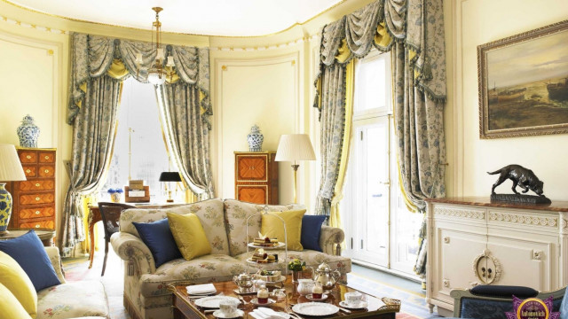 This picture shows a luxurious and modern living room designed by Antonovich Design. The room has a light gray color palette, with light wooden accents on the floor and walls. The furniture pieces are in a subtle cream color with silver and gold accents. There is an ornate fireplace on one side of the room, and two large sectional sofas around a large glass coffee table in the center of the space. The wall is adorned with a large abstract painting and multiple sculptures. The room is completed by the statement ceiling light fixture and curtains that help to create a cozy atmosphere.