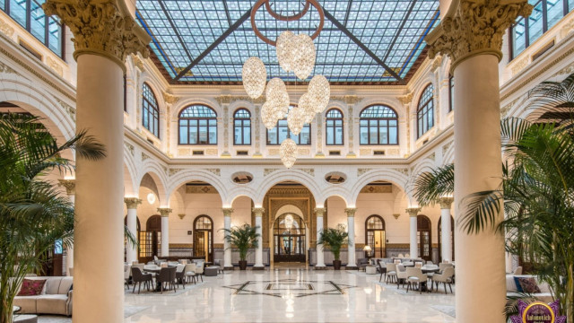 This picture shows an ornately decorated room with modern, Italian-inspired design elements. The walls and ceiling are finished in pale ivory with gold accents throughout, including the crown molding on the ceiling. The furniture is upholstered in a light gray velvet, paired with white marble flooring and dark wood accents. To complete the look, the room is filled with lush plants in pots of various sizes and shapes, adding a touch of nature to the space.