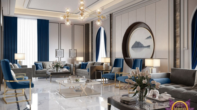 This picture is an interior design concept by Antonovich Design. It shows a modern, contemporary living room with two white leather couches facing each other across a round glass coffee table. The walls are a light beige color with a decorative stone accent wall. There is a fireplace on the wall and two large windows with white curtains. A light grey rug lies across the floor, and there are several potted plants that bring some life and nature into the room.