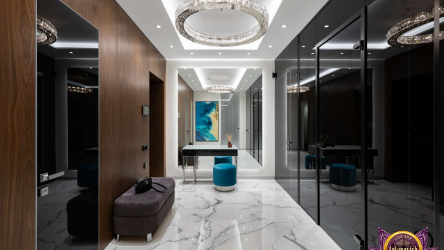 This picture shows a beautifully designed living room, with a luxurious white and grey colour palette. The focal point of the room is a comfortable white armchair, with a white marble table placed in front. To the left is a contemporary glass sofa set, and to the right is a modern wooden display cabinet. An intricate patterned rug covers the floor, while floor-to-ceiling windows on the back wall allow plenty of natural light into the room.