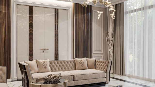 This picture shows a modern living room with a stylish white sofa and two armchairs, both of which are upholstered with a black patterned fabric. The walls, ceiling, and floor have been finished with a light wood veneer and the furniture has been accentuated with gold-coloured accents. There is a decorative rug in the centre of the room and a coffee table in front of the sofa. On the left side of the room, there is a large modern television mounted on the wall and a variety of plants arranged around the furniture.