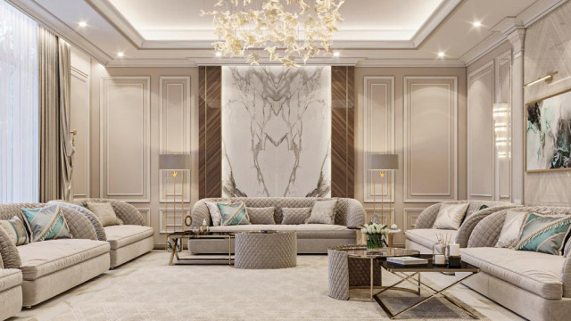 This picture shows a modern, luxurious living room designed by Antonovich Design. It features dark hardwood flooring with a light colored rug, white walls and ceiling, and two large windows on the wall. The furniture consists of a grey velvet fabric sofa with white accents, two black leather ottomans, and several white and gold accent chairs with matching end tables. There is also an art piece above the sofa, with a tall glass vase filled with white flowers adding a touch of sophistication.
