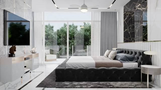 This picture shows a luxurious bedroom featuring a large bed with a metal frame, a cream-colored headboard, and several coordinating pillows and blankets. The walls are painted a soft gray and decorated with wall art, while the floor is carpeted in a plush, light gray shag rug. Other furniture includes two small side tables with lamps, a large mirrored sideboard, and two wingback chairs with ottomans.