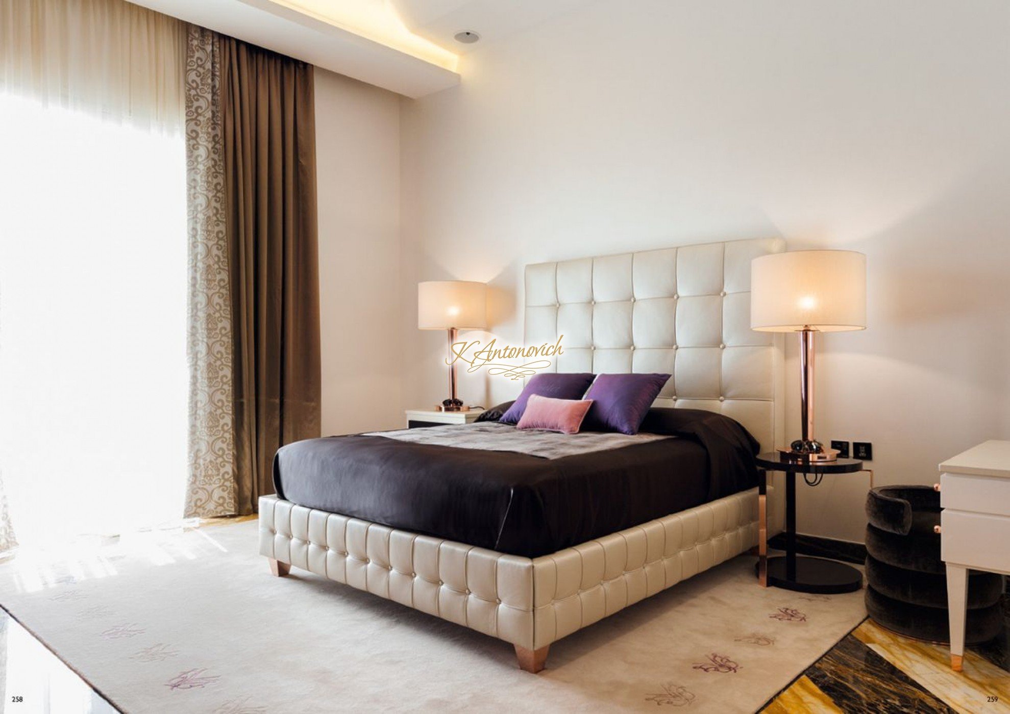 This picture is a photo of a luxurious bedroom interior design. It features a large, upholstered king-size bed with a tufted headboard and gold detailing in the furniture, including the curved bed frame, nightstands, and decorative mirror. The walls are a soft gray color and the drapery frames the views from the tall windows. There is a cream-colored area rug that has both traditional and modern designs, adding an elegant touch to the room. Other decorative elements such as pendant lights, art, and statement pieces complete the look.