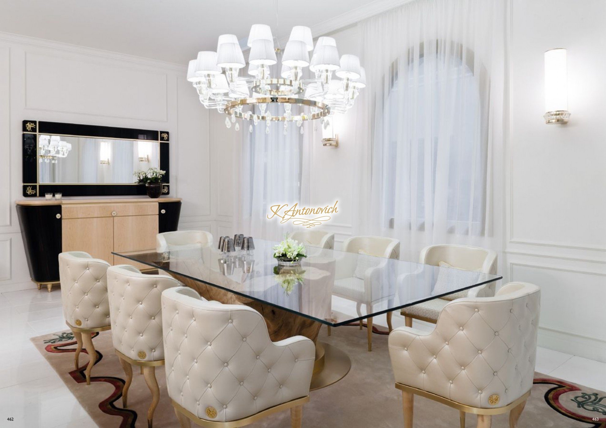 This picture shows a modern living room with a large sectional sofa in a light gray color. There is a round glass-top coffee table in the middle, and two high back chairs in a matching color. The walls are decorated with abstract canvas art pieces, and a large mirror is hung above the sofa. The room has hardwood floors and a large, crystal chandelier hanging from the ceiling.