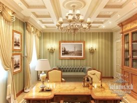 This is a picture of a modern living room. The room features a comfortable grey couch with a cushioned cream footstool in front. The walls and ceiling are painted a light grey and accented by crown molding and a white ceiling fan. Several pieces of stylish decor are found throughout the room, ranging from wall art and a floor rug to fashionable table lamps and a floating wooden shelf.