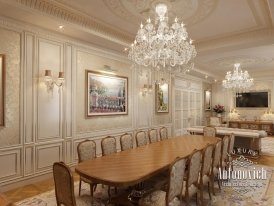 This is a picture of a luxurious interior room. The room features a sophisticated cream and gray color palette, with a gray chaise lounge sofa, two white armchairs, a carved wooden coffee table, and a decorative pink and white vase with a bouquet of flowers. There is an ornately decorated layered chandelier hanging from the ceiling, and a large window overlooking a lush green garden.