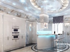 The picture shows a modern white kitchen with sleek marble countertops, custom cabinetry, and stainless steel appliances. The kitchen island is situated in the center of the room and features a built-in sink and stovetop. A breakfast bar lines one side of the kitchen, and there is a large window above the sink for lots of natural light.