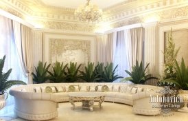 This picture shows an elegant and luxurious sitting area in a grand, ornate interior. It features two comfortable armchairs upholstered in a light fabric, with a matching curved sofa and poufs in the middle. The walls are covered in white wallpaper with elaborate silver accents and a striking golden chandelier hangs from the ceiling. There is also an intricate patterned rug and a glass-top center table.