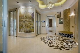 The picture shows a luxurious bathroom, decorated with marble floor and walls, and gold accents. The vanity area features a wood vanity with a double sink, along with additional storage cabinets. Gold fixtures can be seen throughout the bathroom, and a large soaking tub is situated in the background. In the foreground, a white bench can be seen, providing seating near the vanity.