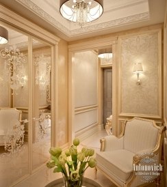 This picture is a rendering of a luxurious entryway designed by Antonovich Design in Dubai. The entryway features a grand marble staircase decorated with an ornate golden railing, with a patterned carpet running up the stairs and down the hall towards the entrance to the room. The walls are decorated with intricate wood paneling and a detailed white trim, and two large wall-mounted mirrors provide additional light and reflection. On either side of the entrance, two marble columns help to frame the space and add to the feeling of grandeur.