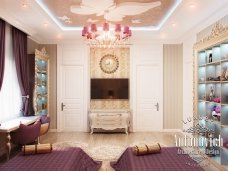 This picture shows an ornate, luxurious living room designed by Antonovich Design in the UAE. The room features white walls with golden trim and details, ornate crown molding and ceiling details, a white and black marble floor, and two deep tan couches with matching chairs and a round coffee table. A deep blue chandelier hangs from the ceiling, and the room is highlighted with various elegant artwork, statues, and accessories.