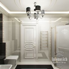 The picture shows a luxurious bathroom in an upscale home. The room has a white tile floor and walls, with a large wooden vanity with two sinks and plenty of counter space. There is a white bathtub with a glass shower door in the corner of the room, and a large window next to it. There are also luxury lighting fixtures and wall sconces, adding to the elegant atmosphere.