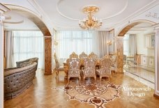 This picture shows a luxurious living room with a stunning crystal chandelier as the main feature. The walls are painted in a beige color, and there are several pieces of gold-plated furniture, such as a sofa, table, and chairs. To add to the opulence, various pieces of art and statues are scattered around the room. A large patterned rug and multiple lamps further enhance the lavish look of the living room.