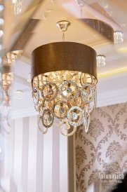 This picture shows a luxurious hallway with a large, ornate chandelier. The walls are a light gray color with a patterned wallpaper on the bottom half. There is a bench with a beige cushion underneath the window, and the floor has a light gray marble tile. The hallway is flanked by two white pillars and the ceiling is decorated with intricate moldings.