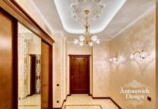 This picture shows a luxurious, modern, and grand entryway. The walls are lined with a light gray wallpaper that has an intricate pattern. On the left wall is a set of tall white columns that support a curved archway. Below the archway is a large round, pink carpet that stretches all the way to the two marble stairs. On the right side is a beautiful chandelier that hangs from the ceiling. Above the stairs is a set of two windows with gold trim. At the end of the stairs is an elegant door with gold handrails leading up to it.