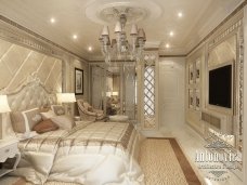 This picture shows a luxurious and opulent bedroom design. The room features gold accents and a rich velvet texture in the bedding and the furniture, creating an air of sophistication. There are classic wall sconces to add a touch of glamour, and a large bed with ornate headboard and curtains. A crystal chandelier hangs from the ceiling and provides a hint of elegance and extravagance.