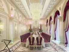 The picture shows a luxurious living room designed with a white and gold color palette. The room features an intricately carved modern sofa with matching armchairs, a white marble coffee table, and an elegant glass chandelier hanging from the high ceiling. The walls are painted in an ivory color and decorated with ornate fabric panels. Gold mirrors and framed artwork provide additional visual interest.