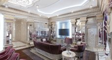 This picture shows the interior of a luxurious and modern living room. The walls have been painted in a light grey tone and feature several large, framed artworks. The centerpiece of the room is a white leather couch with multiple colorful accent pillows. A round, glass-top coffee table sits in the center of the room, surrounded by two matching armchairs. The floor features a light-colored area rug that adds subtle warmth to the space. Additional decorative elements, such as a hanging fixture, house plants, and floor lamps, complete the elegant and modern look of this room.