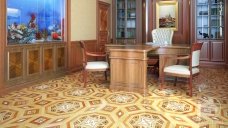 This picture shows an ornate living room. It features a light-colored wooden floor with a large plush white rug in the center, two ivory couches with bright blue pillows, two armchairs upholstered in matching blue fabric, a round glass coffee table, and a round wooden side table. The walls are lined with built-in shelves and drawers, and there are several pieces of artwork displayed on them. A chandelier hangs from the ceiling, providing soft illumination throughout the space.